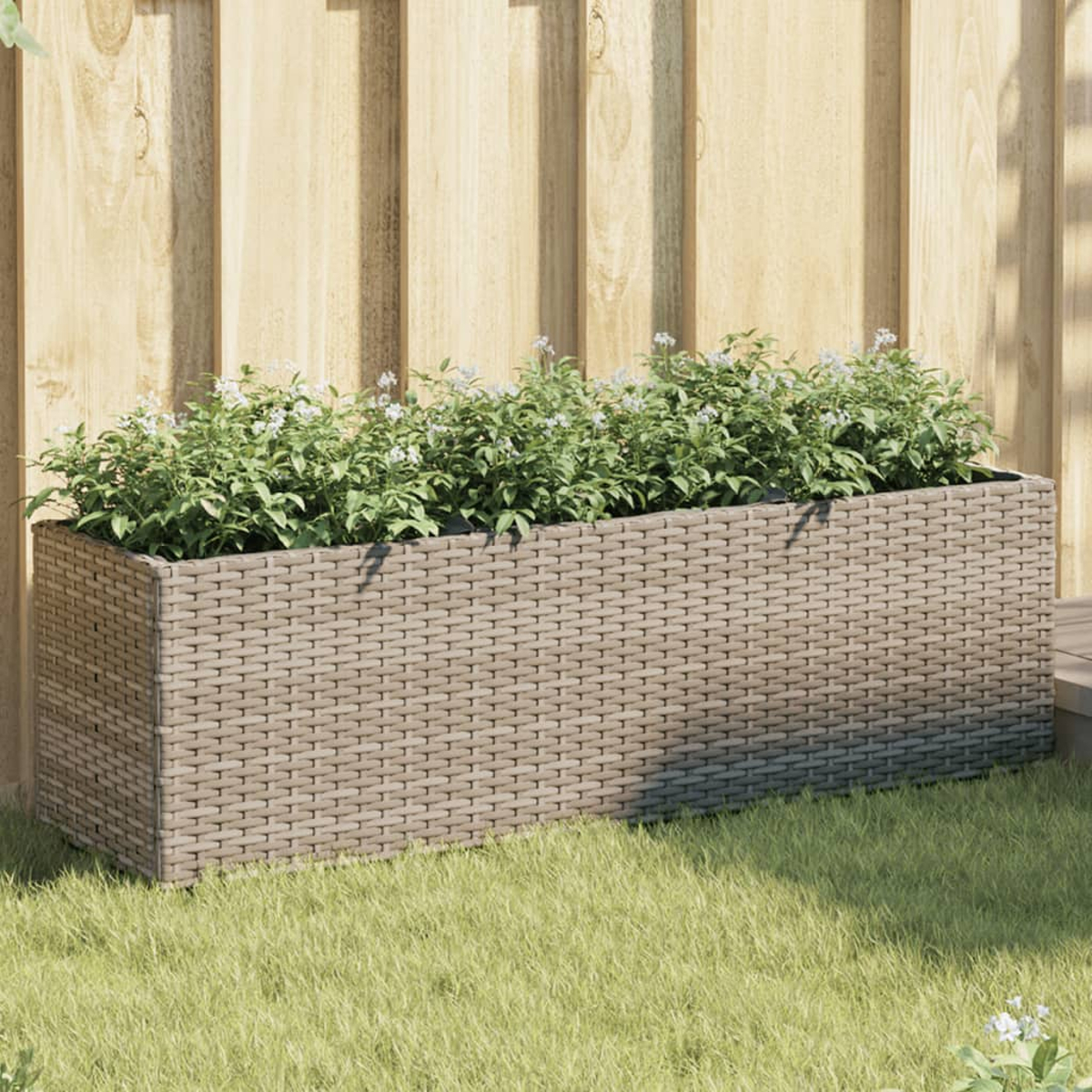Una fioriera rettangolare in vimini tortora con piante verdi e fiori bianchi si trova sull'erba davanti a una recinzione di legno.