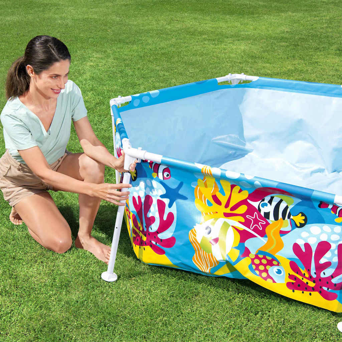 Una donna monta una piscina gonfiabile con decorazioni di pesci e coralli sull'erba.