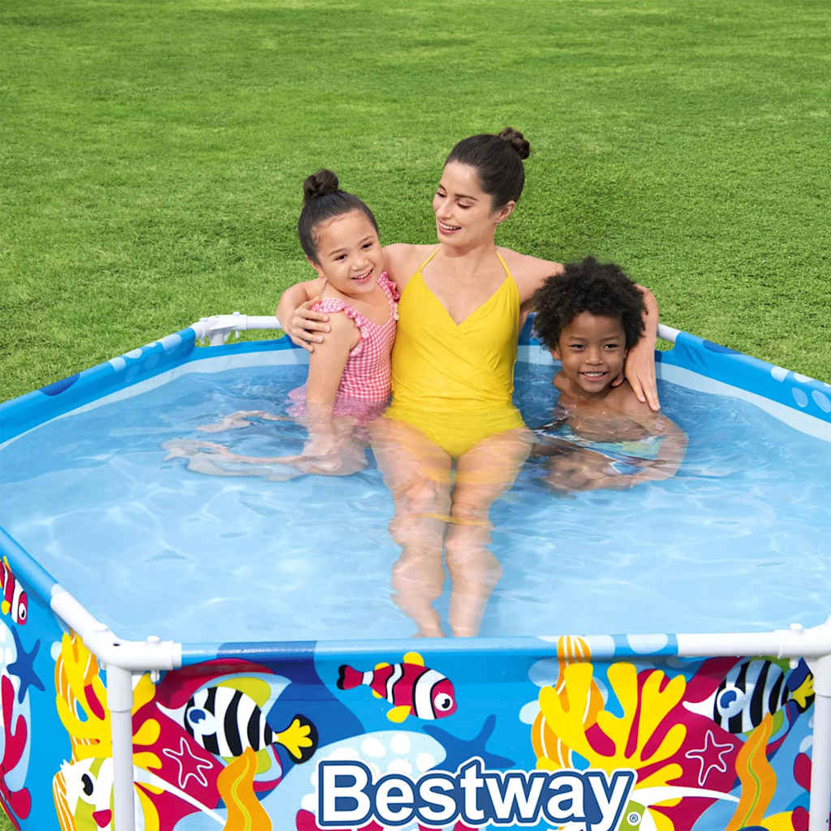 Una donna e due bambini sguazzano in una piscina gonfiabile con disegni di pesci in un giardino.