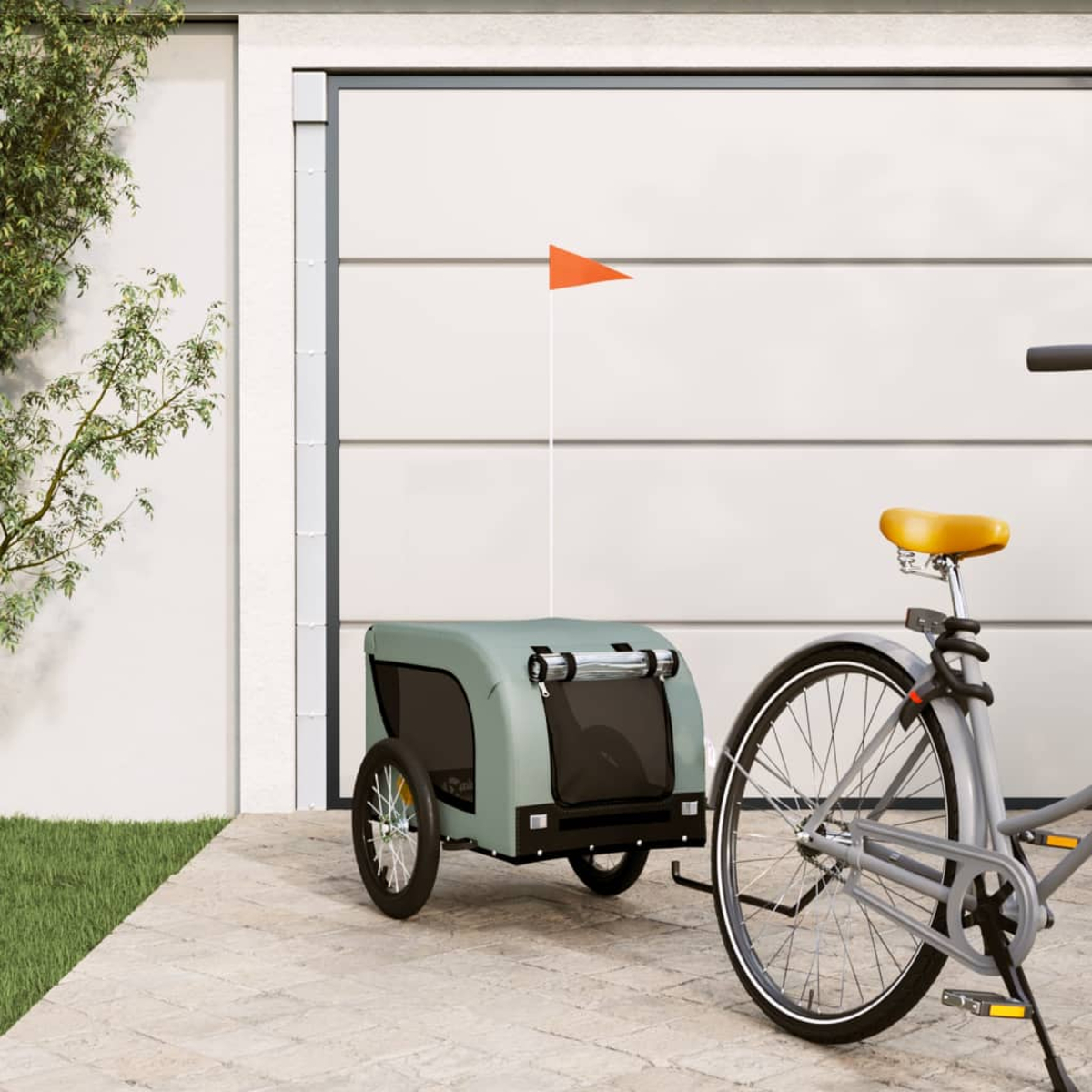 Ein graues Fahrrad mit einem grünen Anhänger und einer orangefarbenen Fahne vor einem Garagentor.