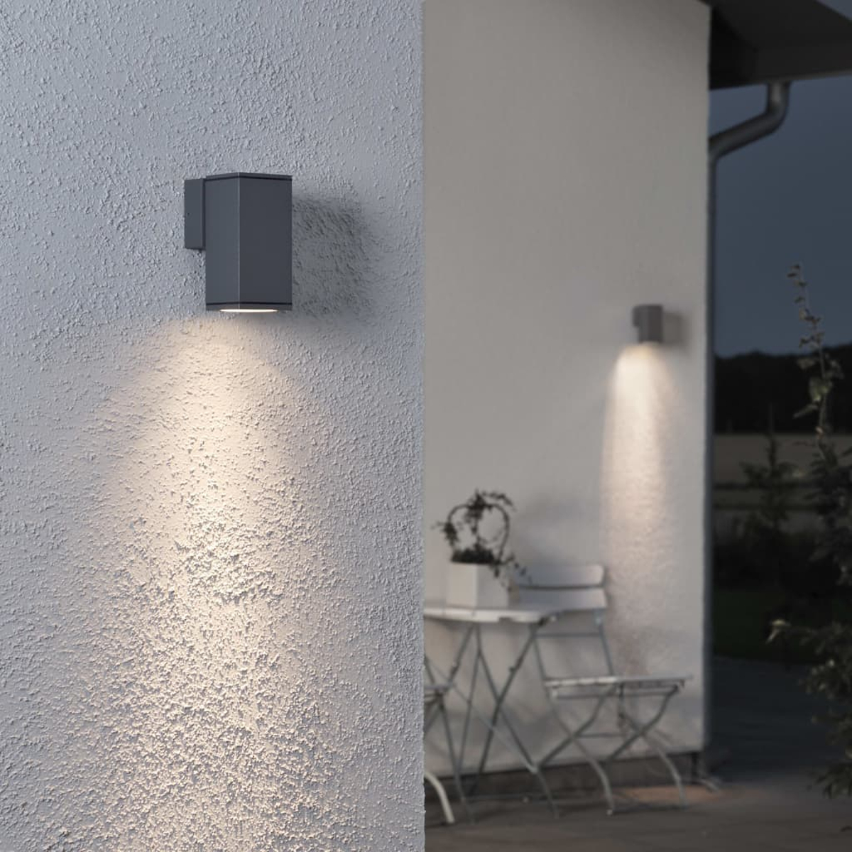 Grauer Außenwandleuchter beleuchtet weiße Wand und Sitzgruppe.