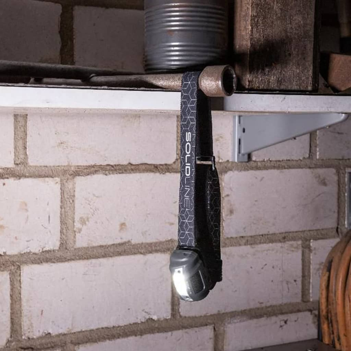 Eine Stirnlampe mit gemustertem Gurt hängt an einem Regal, ihr Licht ist an.