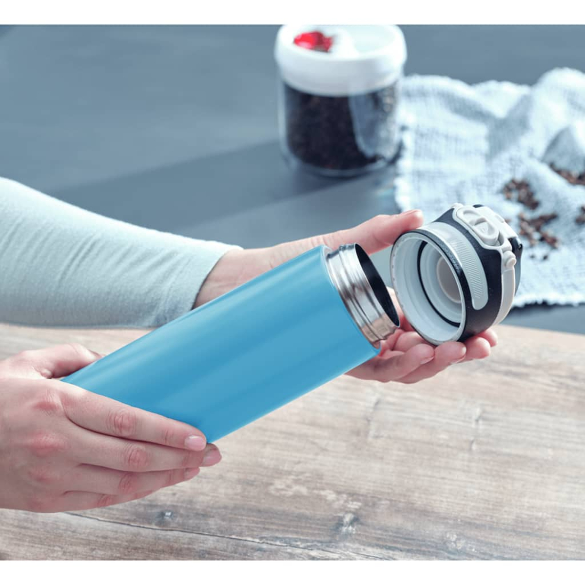 Eine Person öffnet einen blauen Thermobecher mit einem schwarzen Deckel.