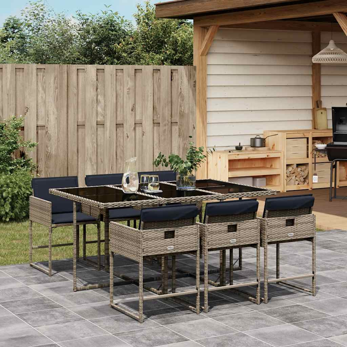 Ein Esstisch aus Rattan mit sechs Hockern steht auf einer gefliesten Terrasse neben einem Grill und Holzlagern.