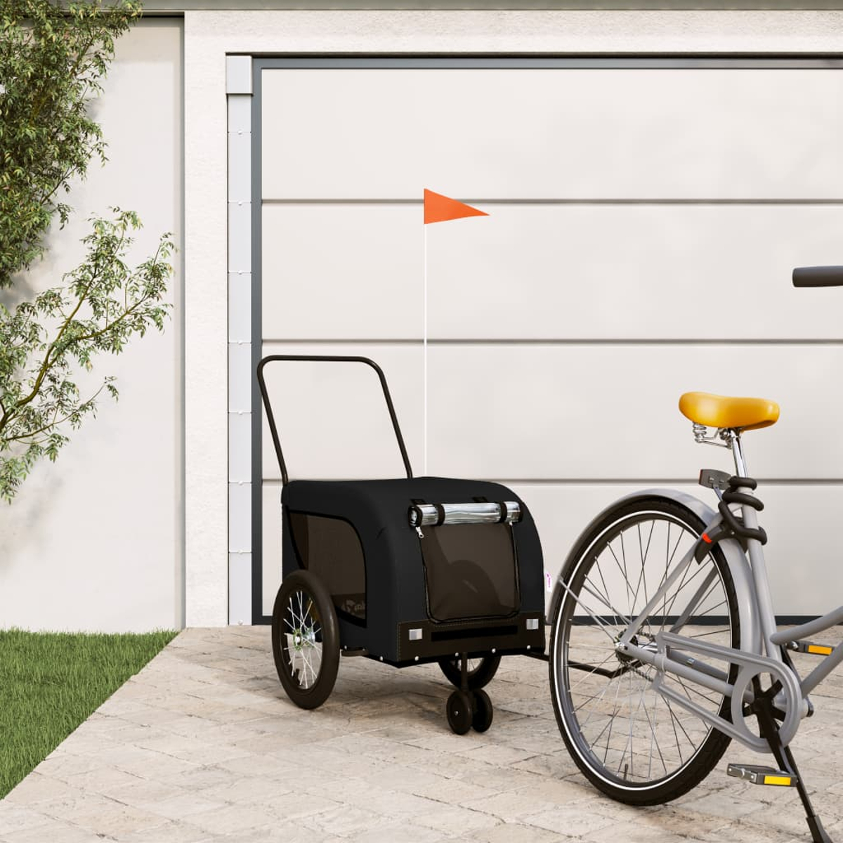 Ein graues Fahrrad ist an einen schwarzen Haustieranhänger mit orangefarbener Flagge angekoppelt, geparkt vor einer Garage.