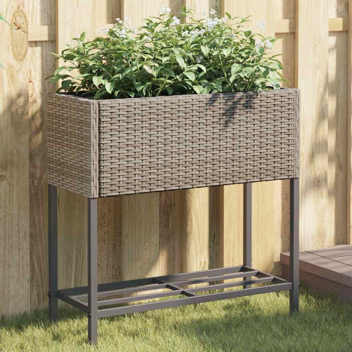 Una fioriera rialzata effetto rattan con supporto metallico si trova sull'erba davanti a una recinzione di legno.