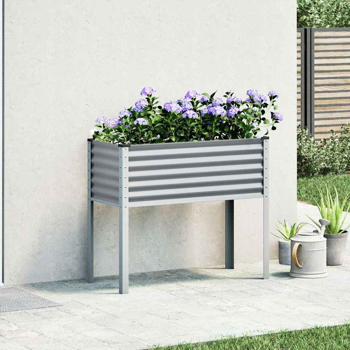 Una fioriera rialzata in metallo grigio con fiori viola si trova su un patio pavimentato vicino a un muro.