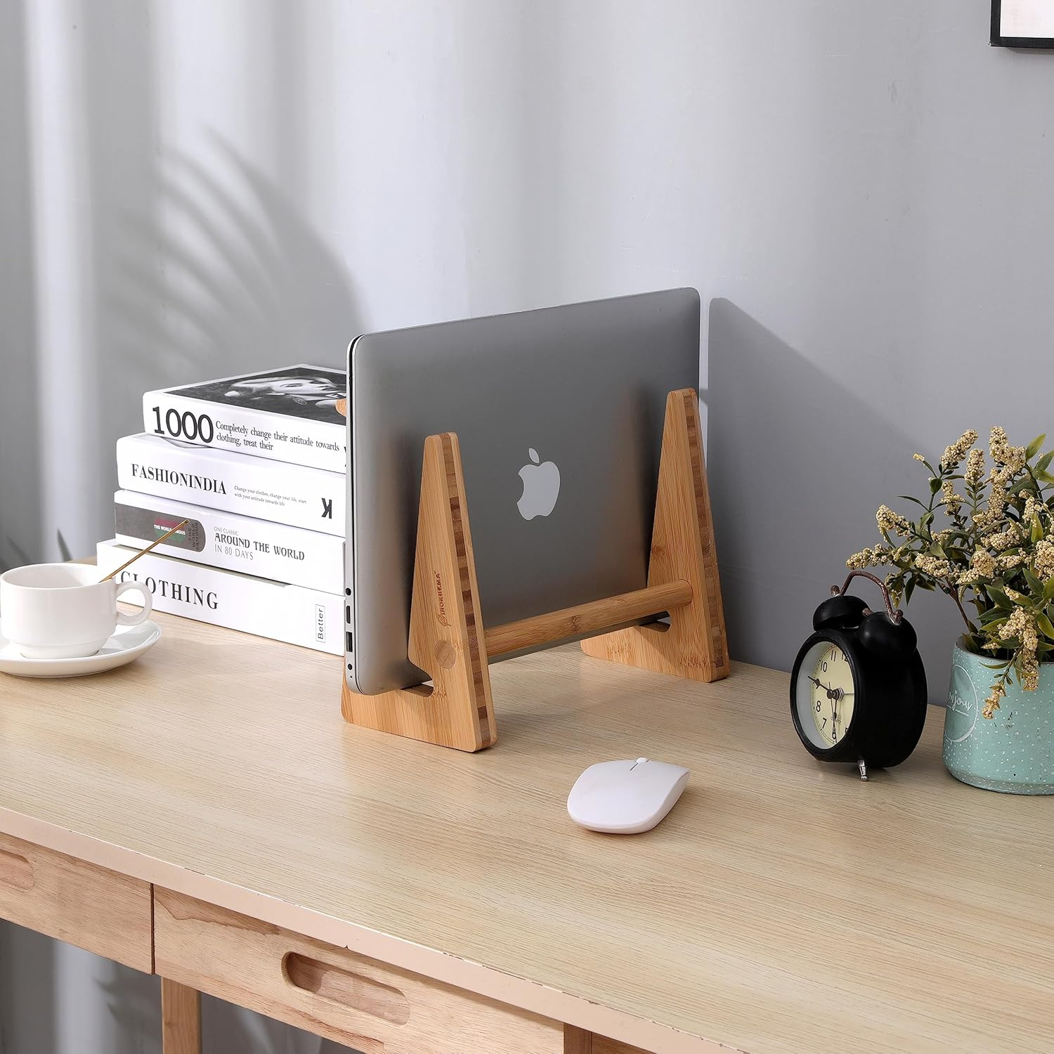 Schreibtisch mit Laptop auf Ständer, Büchern, Tasse, Maus, Wecker und Pflanze.