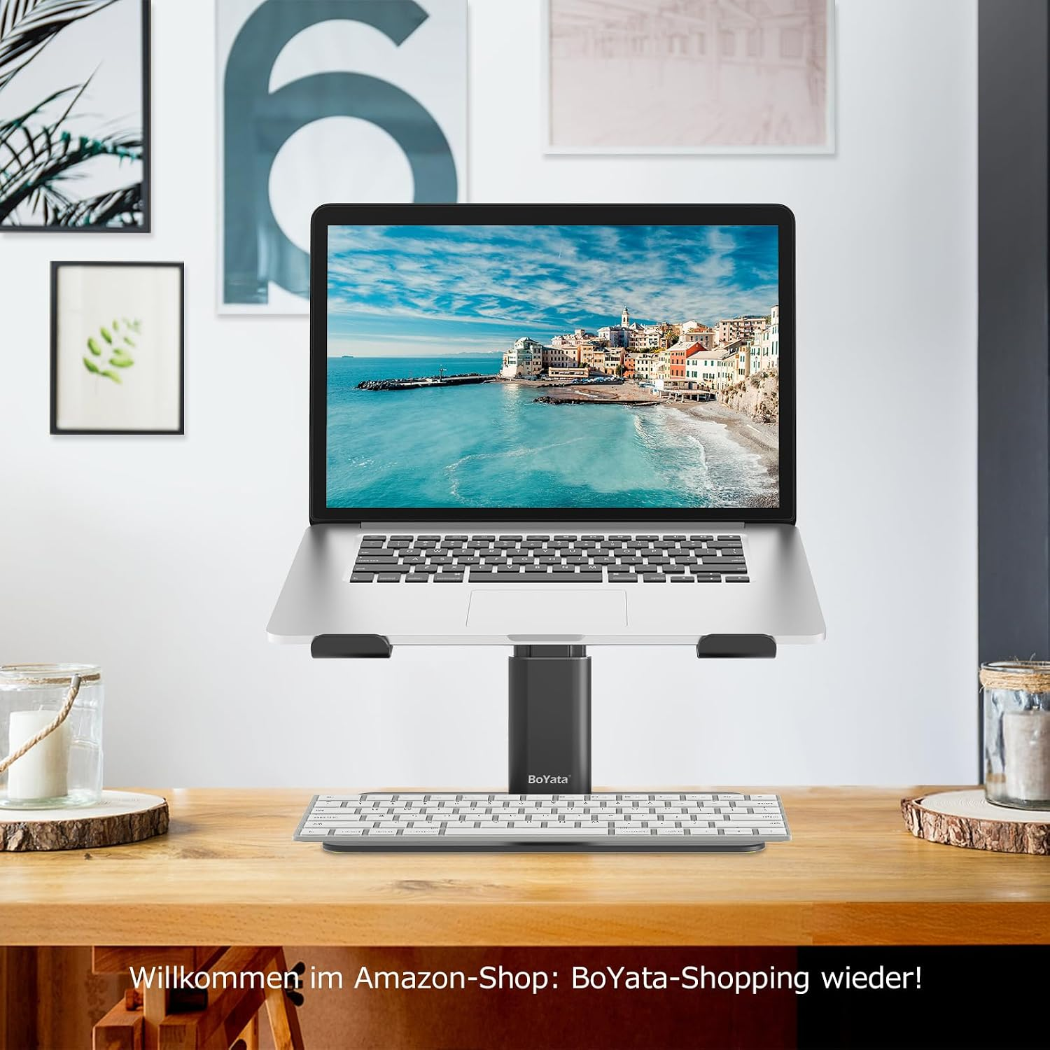 Ein Laptop auf einem Ständer mit einer Tastatur auf einem Holztisch. Im Hintergrund Bilder.