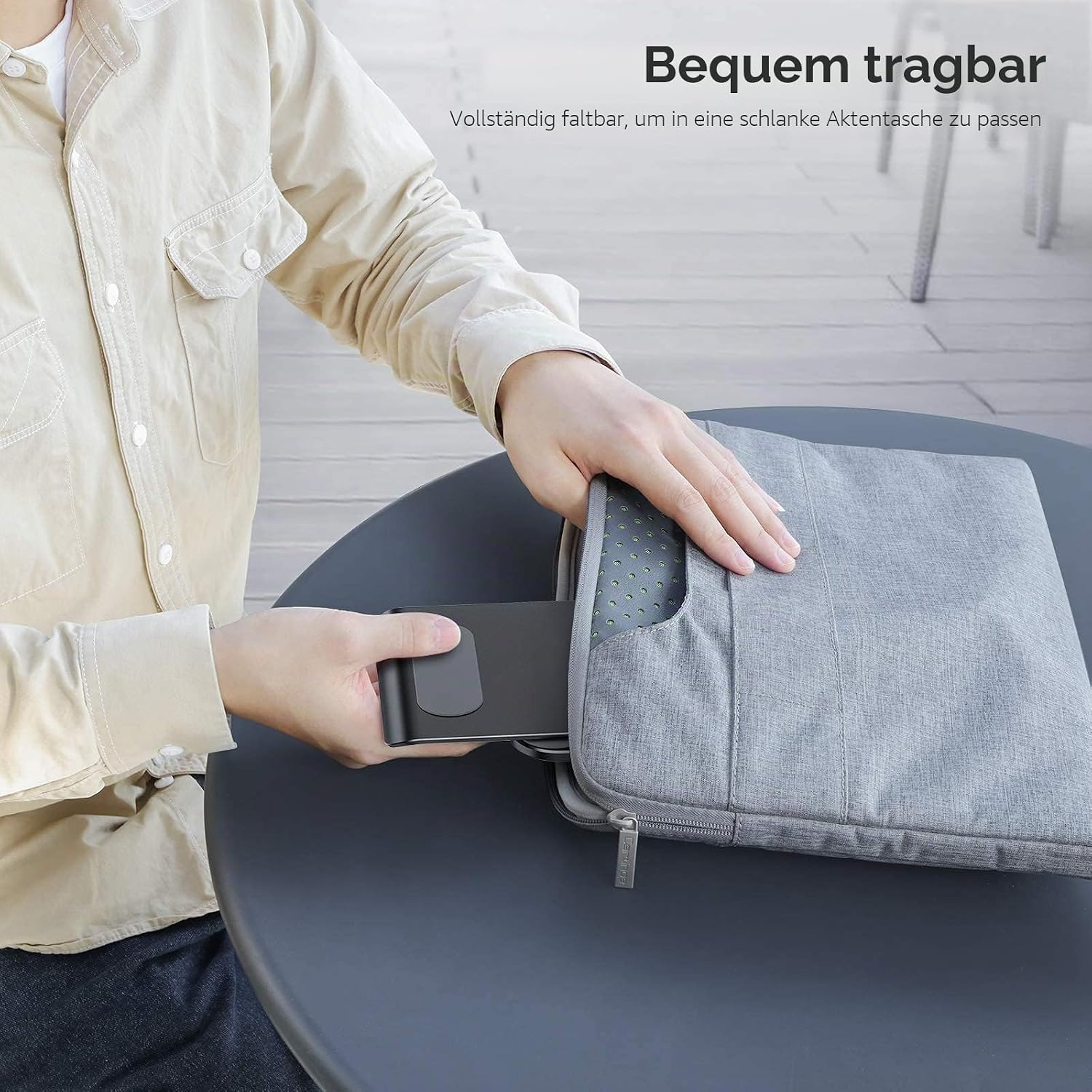 Mann steckt Laptop in graue Tasche. Tisch und Stuhl im Hintergrund.