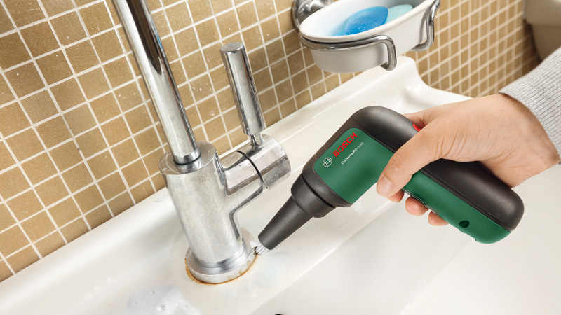 Hand hält Bosch Universalbürste zur Reinigung eines Wasserhahns in einem Waschbecken.