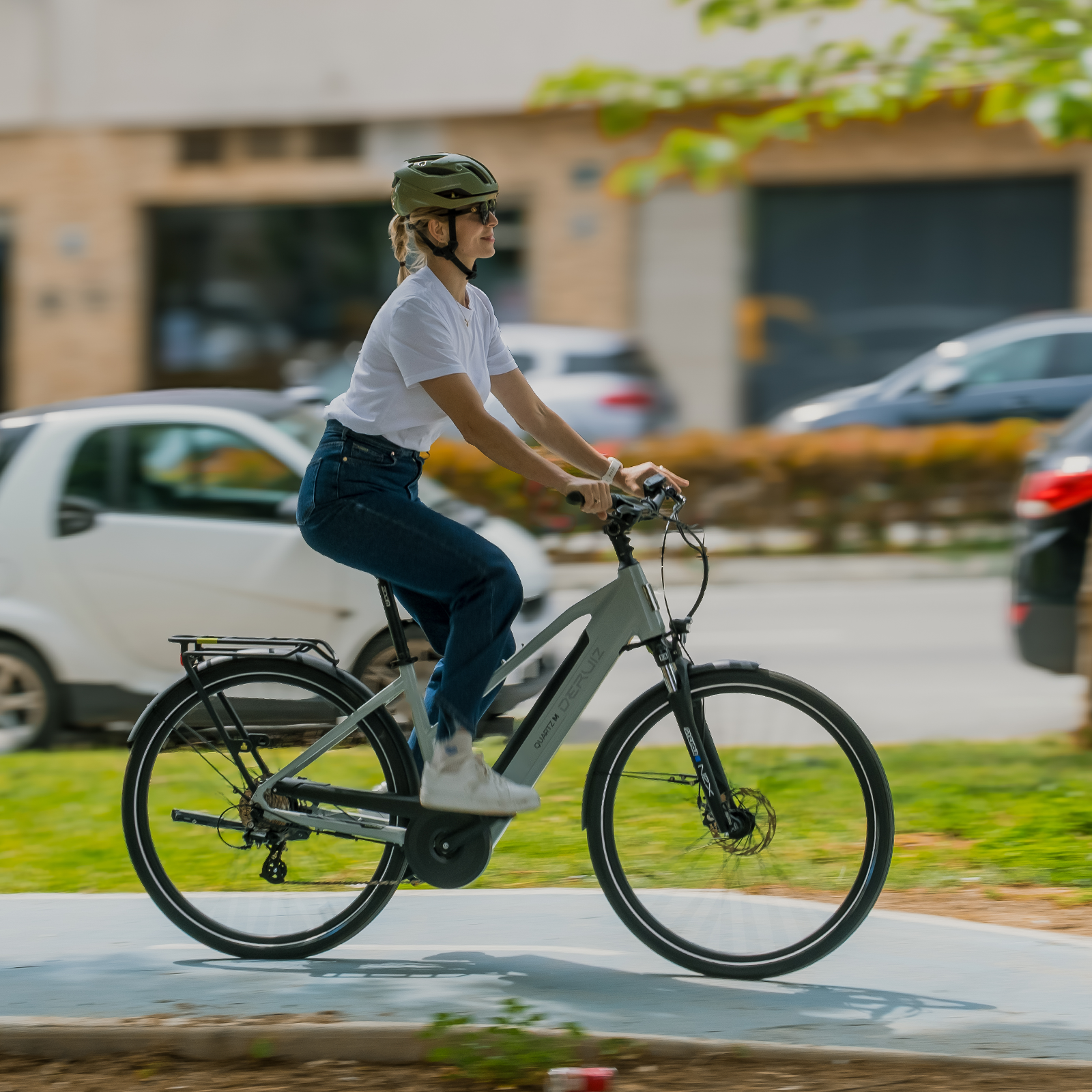 Eine Frau mit Helm fährt ein graues Fahrrad auf einem Weg, Autos im Hintergrund.