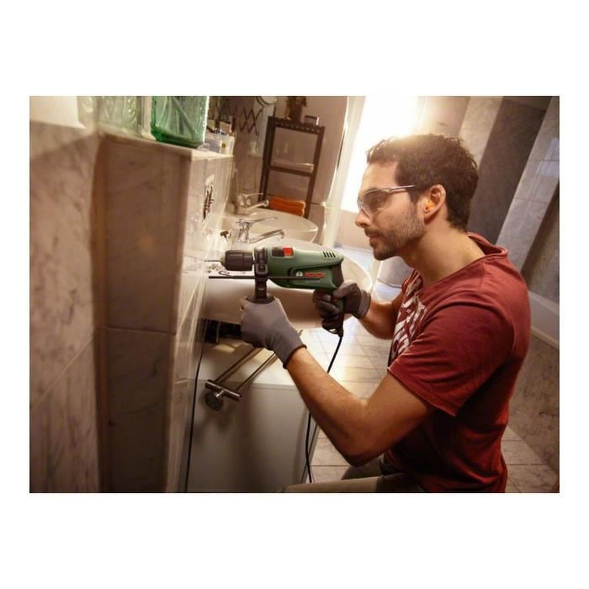 Un hombre taladra en un baño con gafas de seguridad y guantes con un taladro.