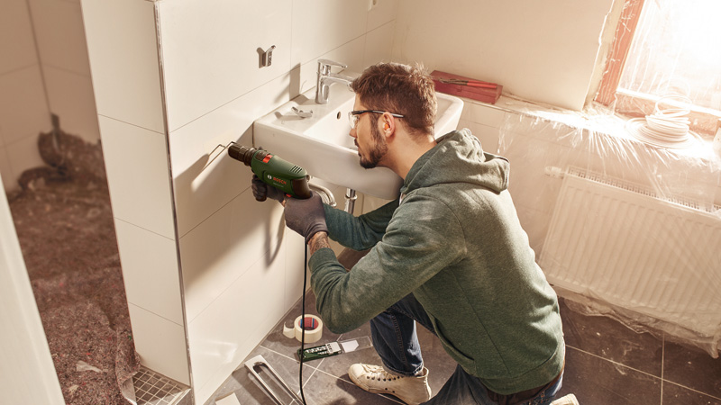 Hombre taladrando pared junto a lavabo, baño en renovación.