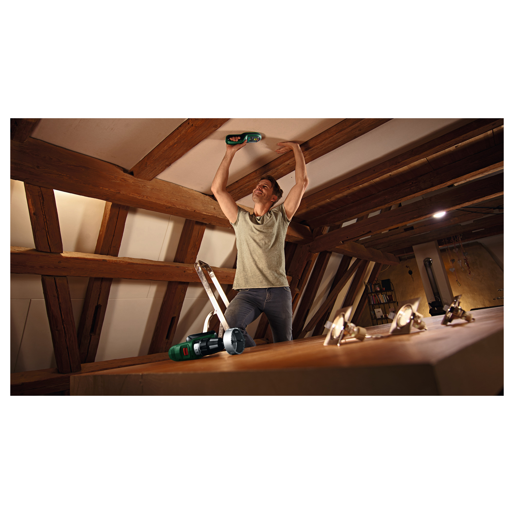 Ein Mann steht auf einer Leiter in einem Dachgeschoss mit Holzbalken und nutzt ein grünes Messgerät an der Decke.