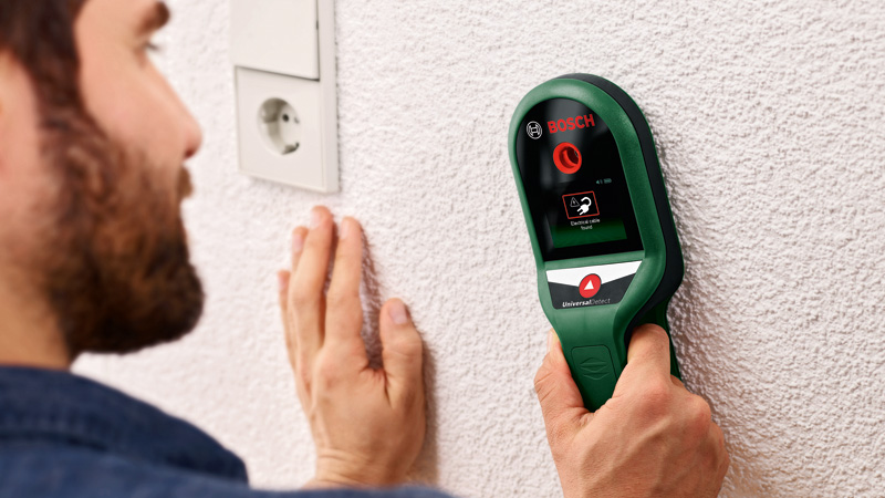 Ein Mann nutzt einen grünen Bosch Ortungsgerät an einer weißen Wand in der Nähe einer Steckdose.