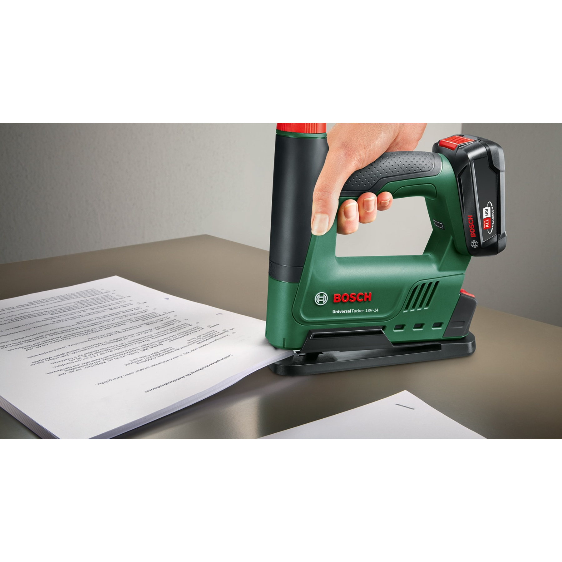 Eine Hand hält einen grünen Bosch Akku-Tacker, der auf Papierblätter auf einem Holztisch gerichtet ist.