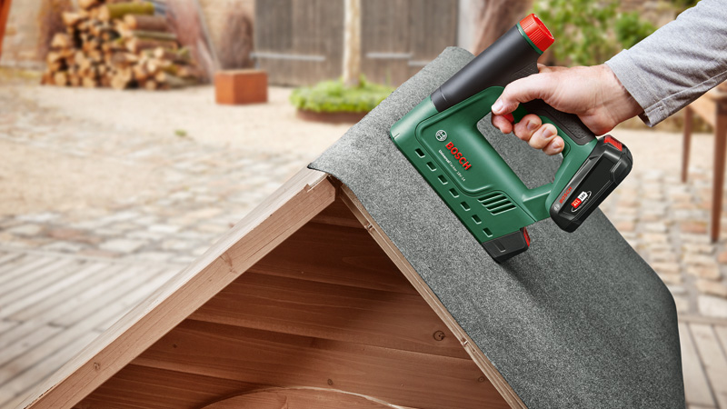 Eine Hand benutzt einen grünen Bosch Akku-Tacker, um Dachpappe auf einem Holzdach zu befestigen.