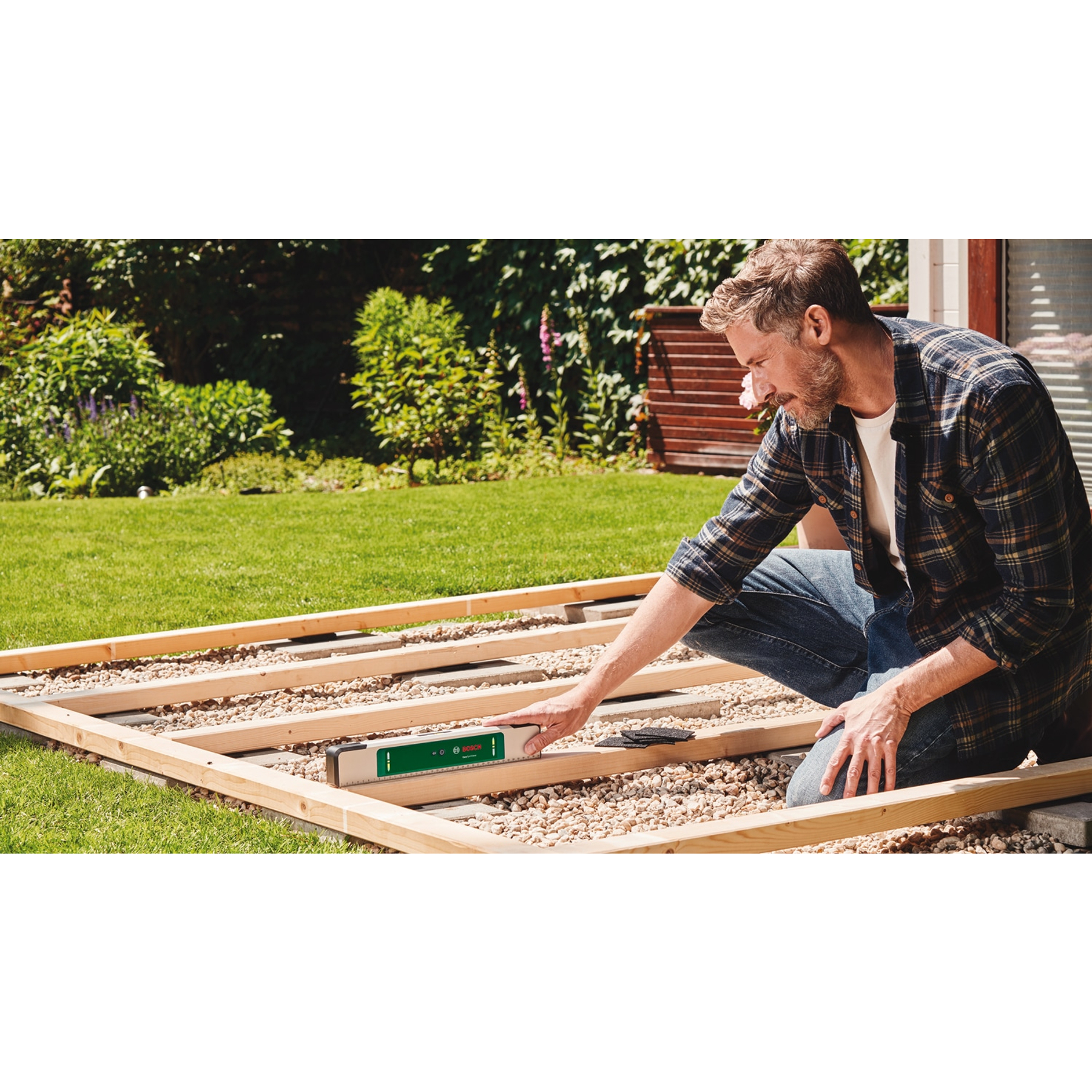 Ein Mann im Karohemd benutzt eine Wasserwaage an einem Holzrahmen über Kies in einem sonnigen Garten.