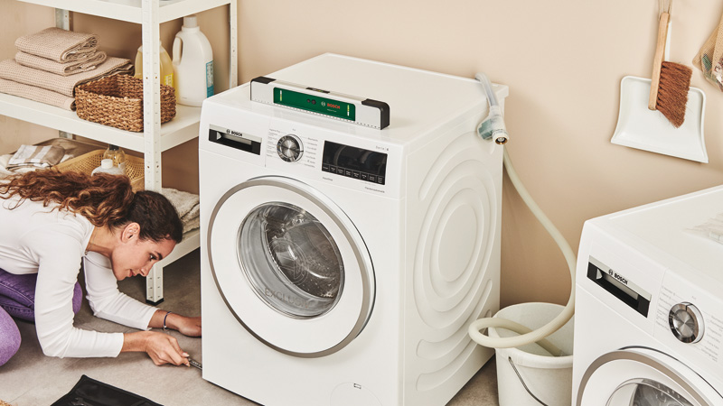 Eine Frau adjusts the leveling foot of a white washing machine with a spirit level on top.
