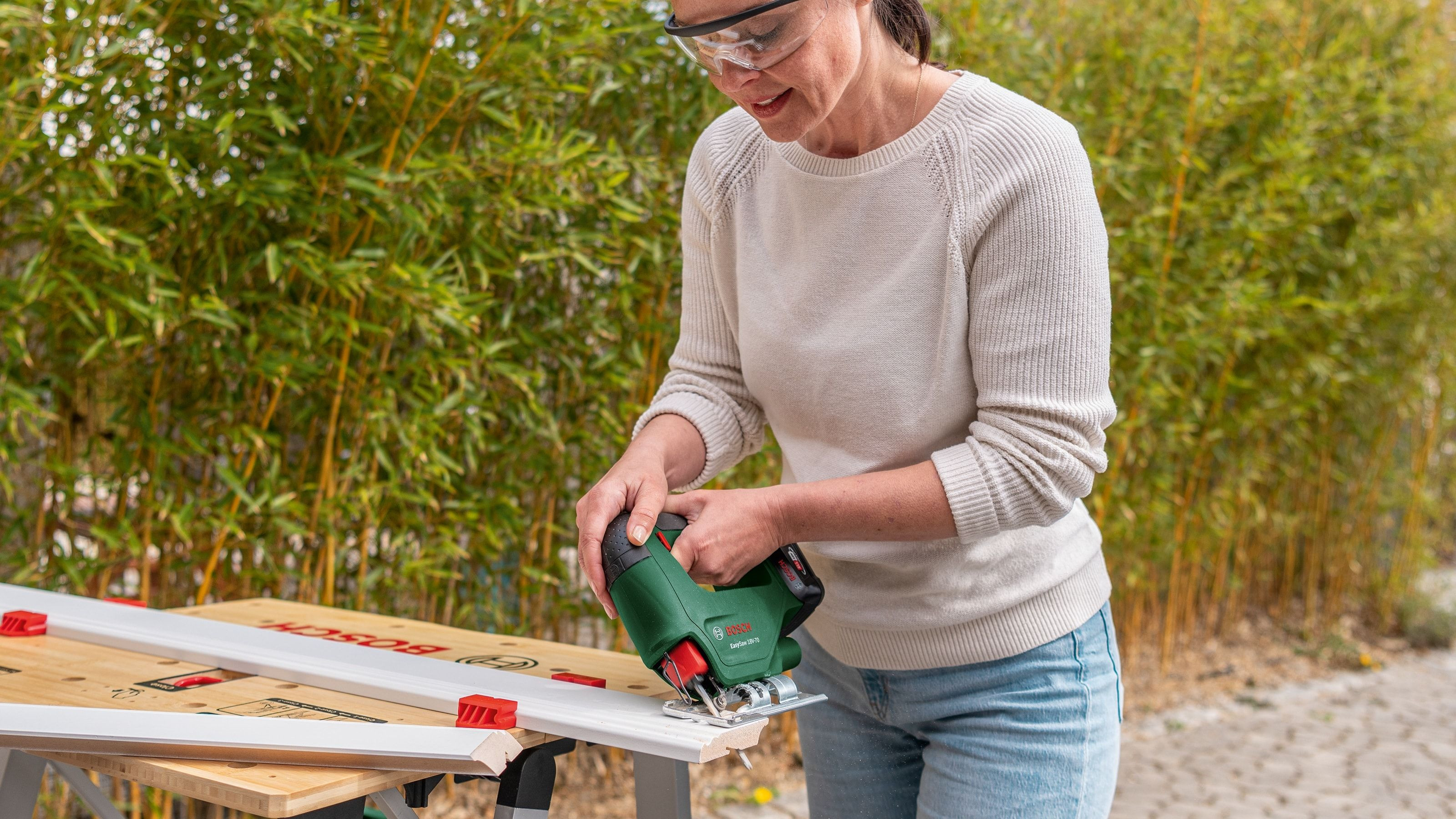 Eine Frau in Schutzzbrille schneidet draußen eine Holzleiste mit einer grünen Stichsäge.