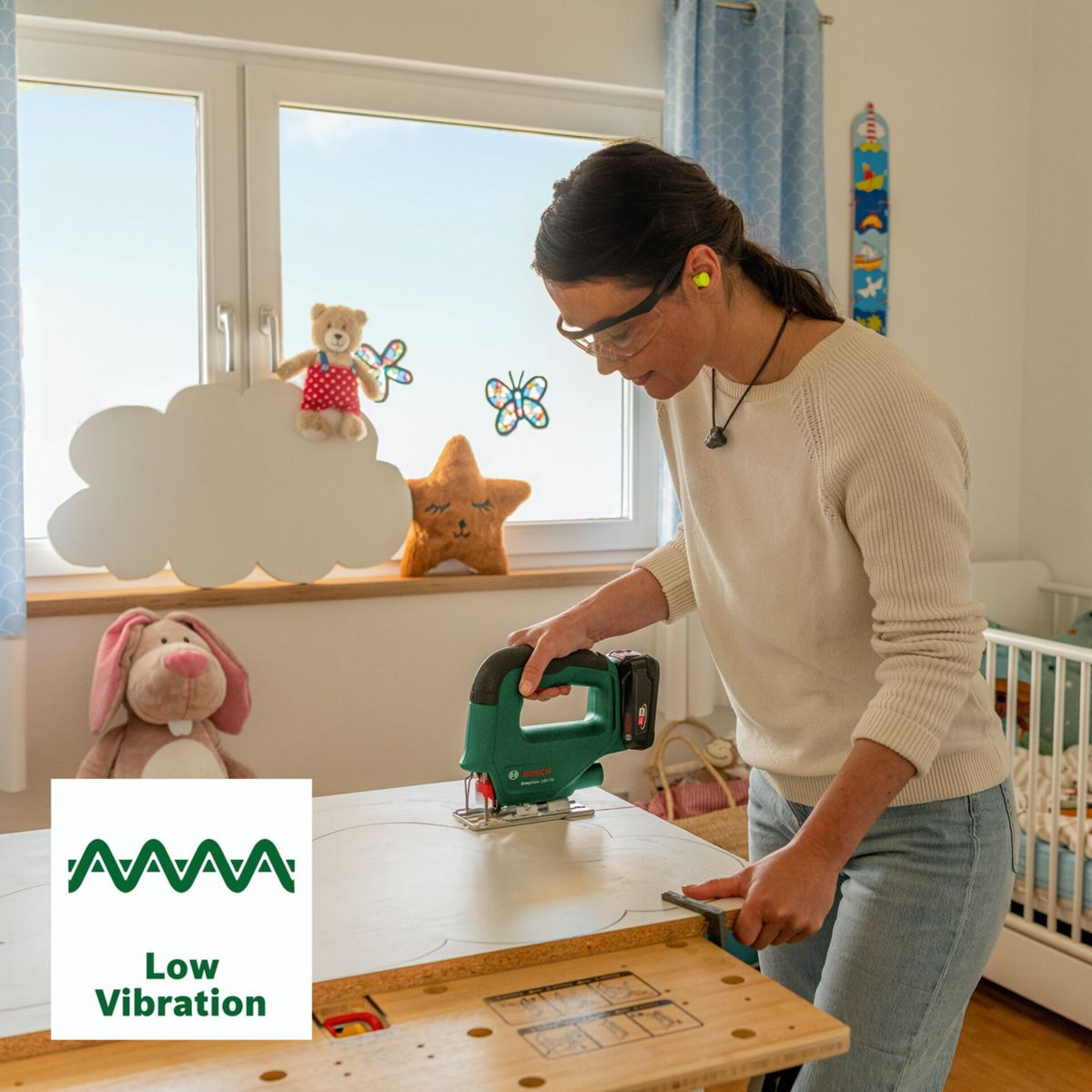 Eine Frau schneidet mit einer grünen Stichsäge eine Holzplatte zu; im Hintergrund ein Kinderzimmer.
