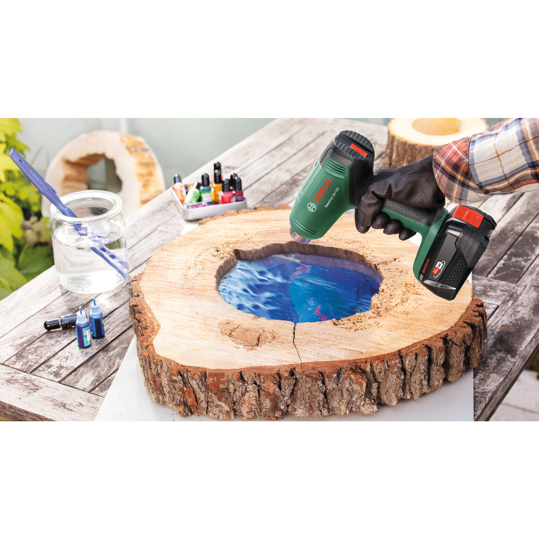 Eine Hand mit Heißluftpistole bearbeitet blaues Epoxidharz in einer Holzscheibe auf einem Tisch.