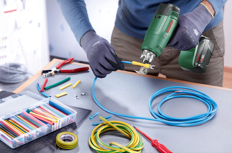 Person arbeitet an einem Tisch und benutzt ein grünes Elektrowerkzeug, um einen gelben Kabelschuh an ein blaues Kabel zu crimpen.