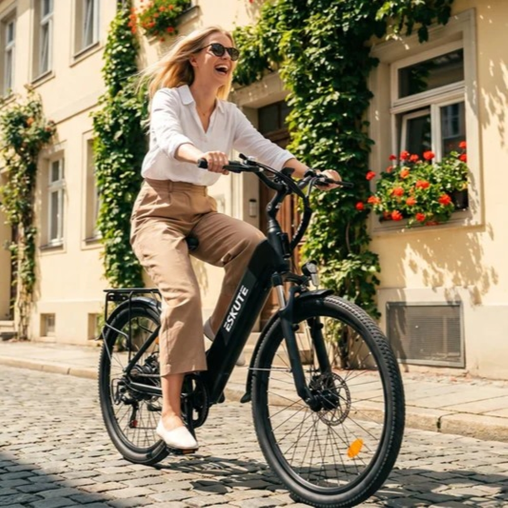 Una donna bionda sorridente in camicia bianca e pantaloni beige guida una e-bike nera.