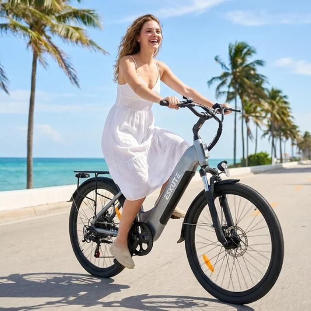 Una mujer sonriente con vestido blanco monta una bicicleta eléctrica gris por una carretera costera con palmeras.