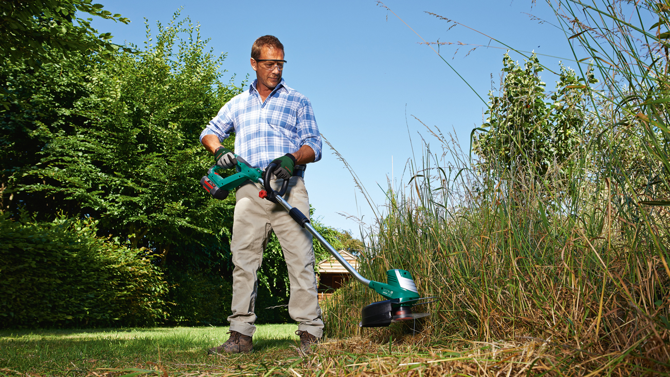 Ein Mann im karierten Hemd schneidet hohes Gras mit einem Rasentrimmer in einem sonnigen Garten.