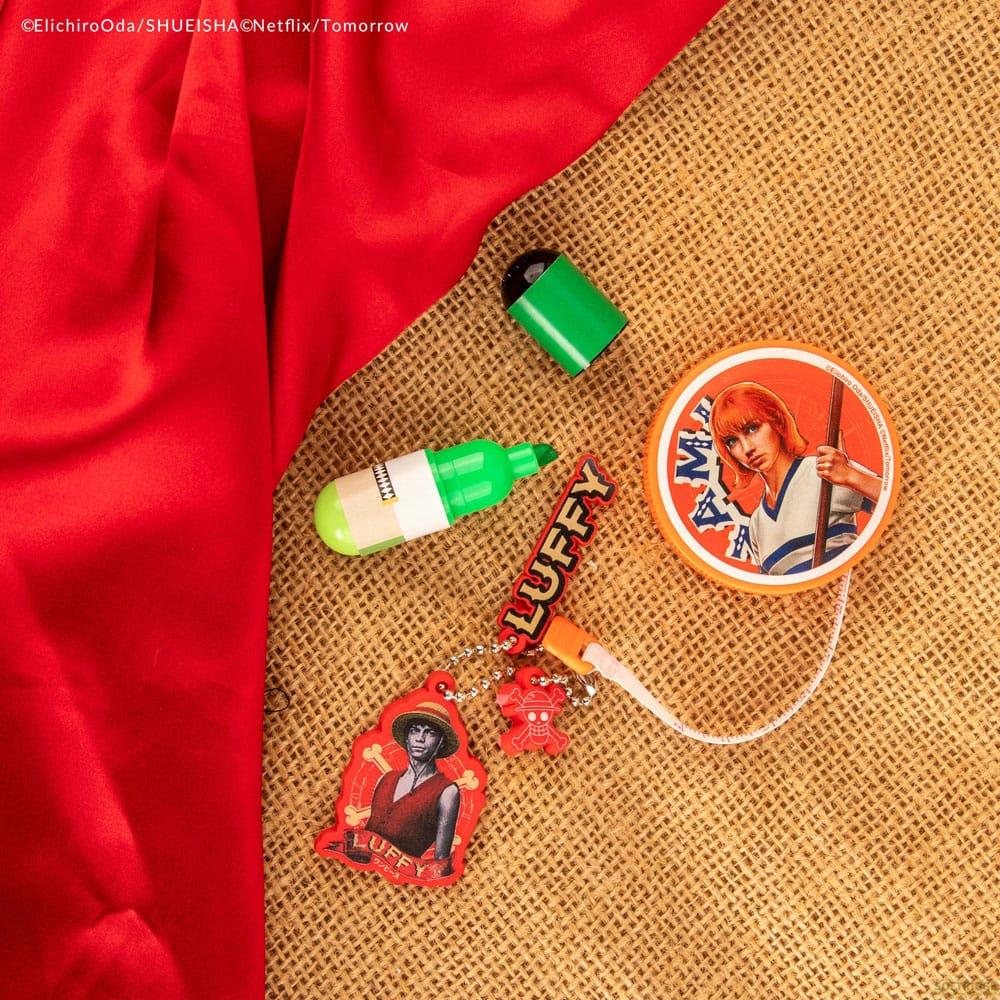 Merchandising de One Piece sobre arpillera: tela roja, llavero de Luffy, pin de Nami y rotuladores verdes.