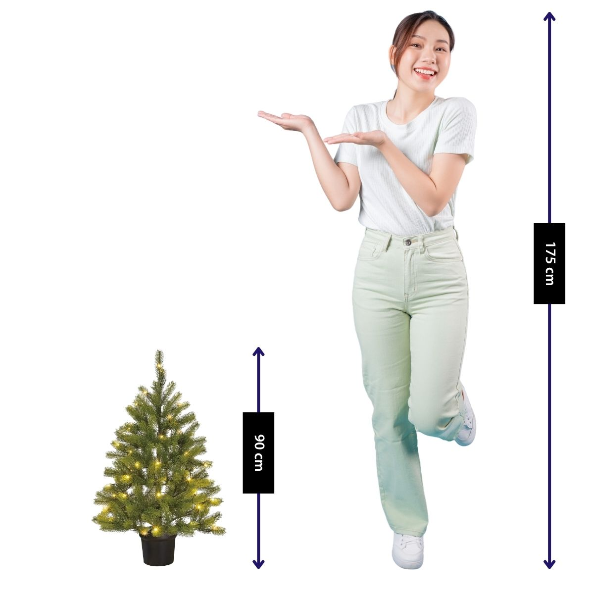 A smiling woman in a white shirt and light pants stands next to a small, lit Christmas tree.