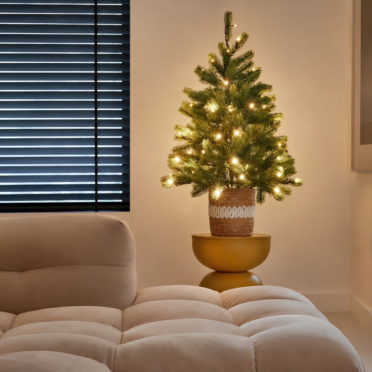 A lit Christmas tree in a woven pot sits on a yellow stool near a beige sofa and a window with blinds.