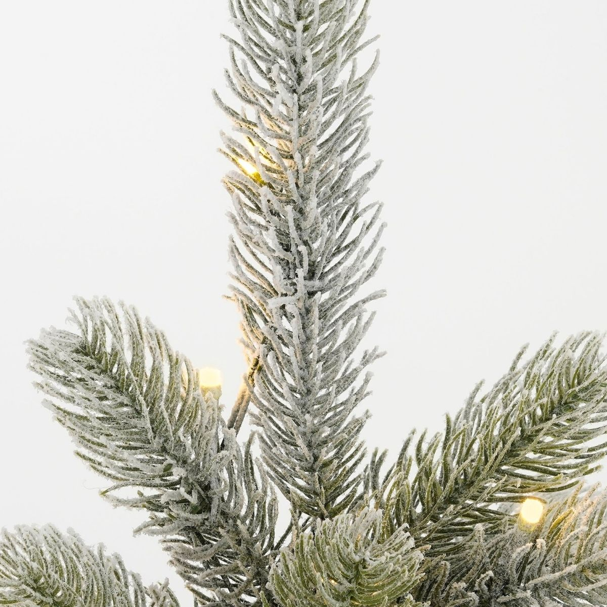 Snow-covered artificial pine tree branches with warm white fairy lights against a plain white background.