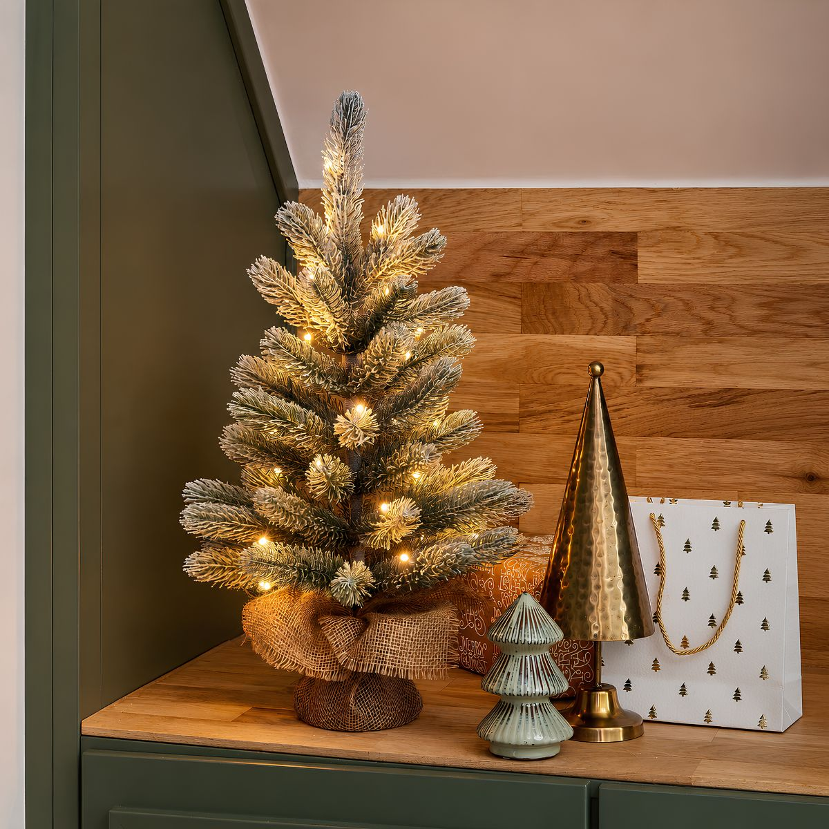 A lit, flocked miniature Christmas tree sits on a wooden shelf next to gold and ceramic decor.