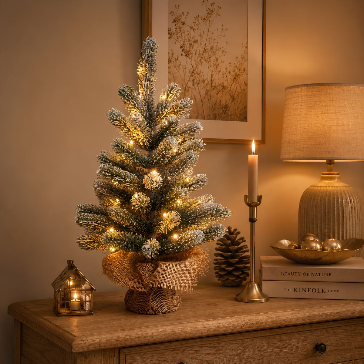 A small snow-dusted Christmas tree with lights sits on a wooden table beside books, a lamp, and a candle.