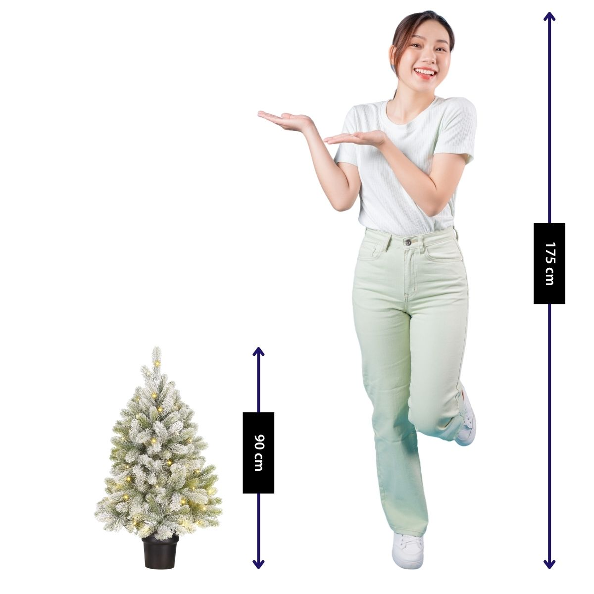 A smiling woman gestures toward a 90 cm snowy mini Christmas tree next to her.