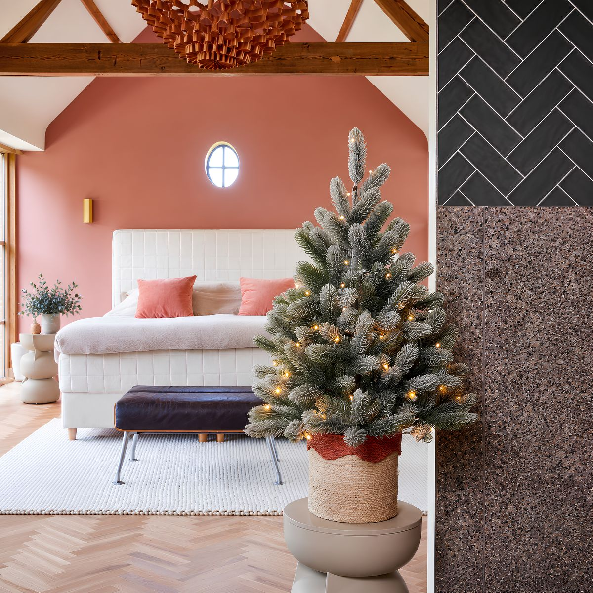 Lit small Christmas tree in a pot in front of a bedroom with pink wall and white bed.