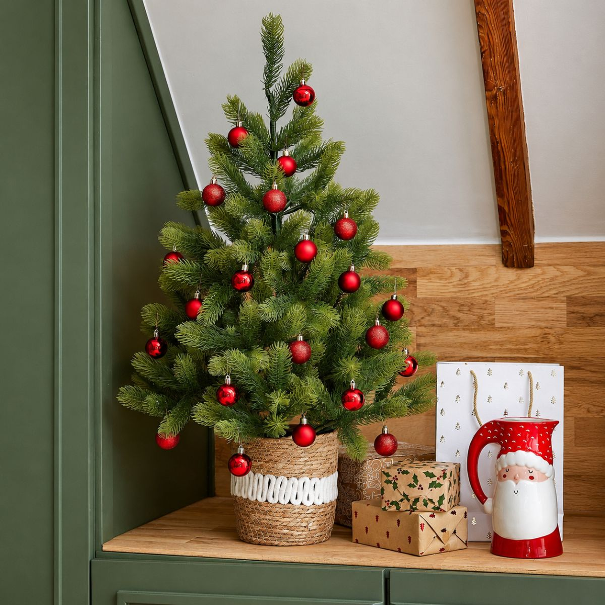 A small Christmas tree in a woven basket, decorated with red baubles, stands next to wrapped gifts and a Santa Claus jug.