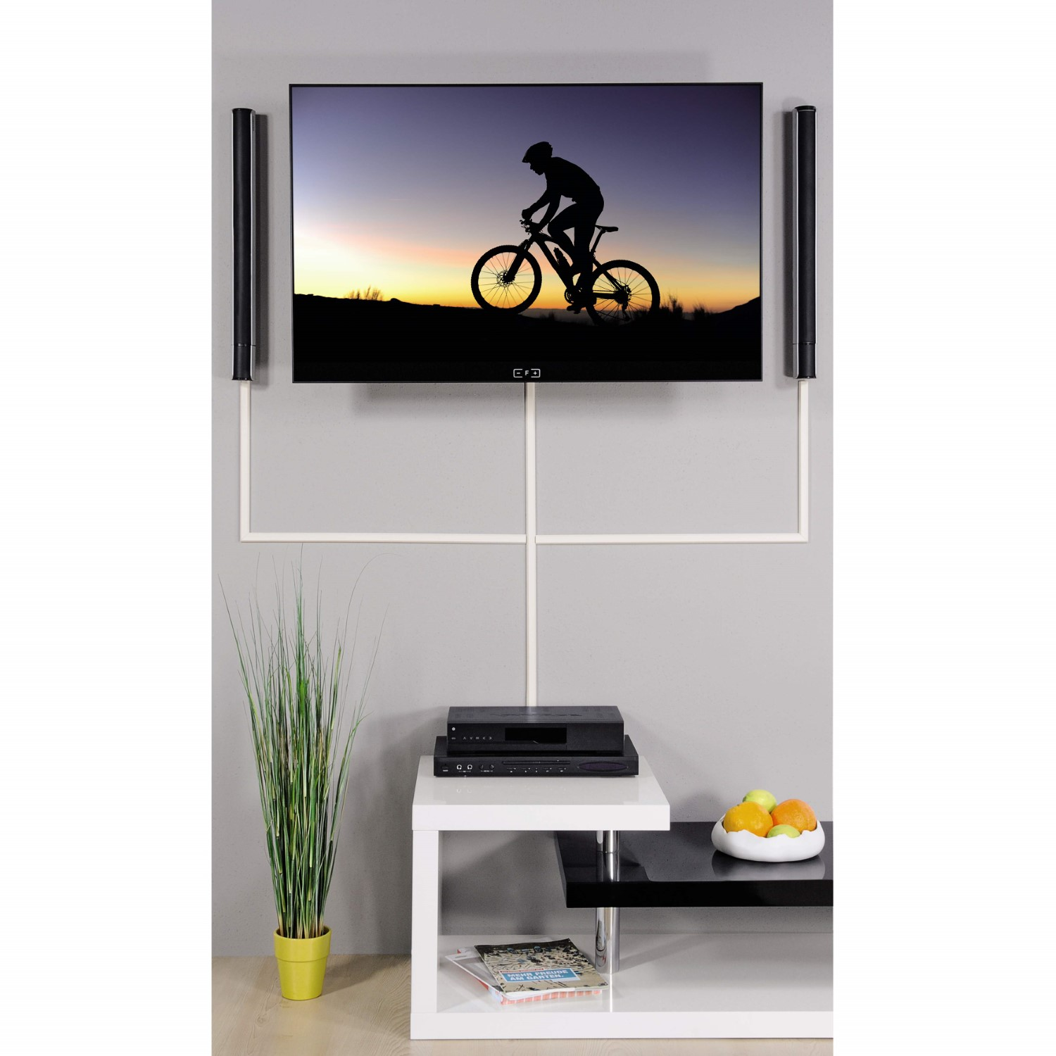 Wandfernseher mit Fahrradmotiv, Lautsprechern, Kabelkanal und TV-Möbel mit Pflanze und Obst.