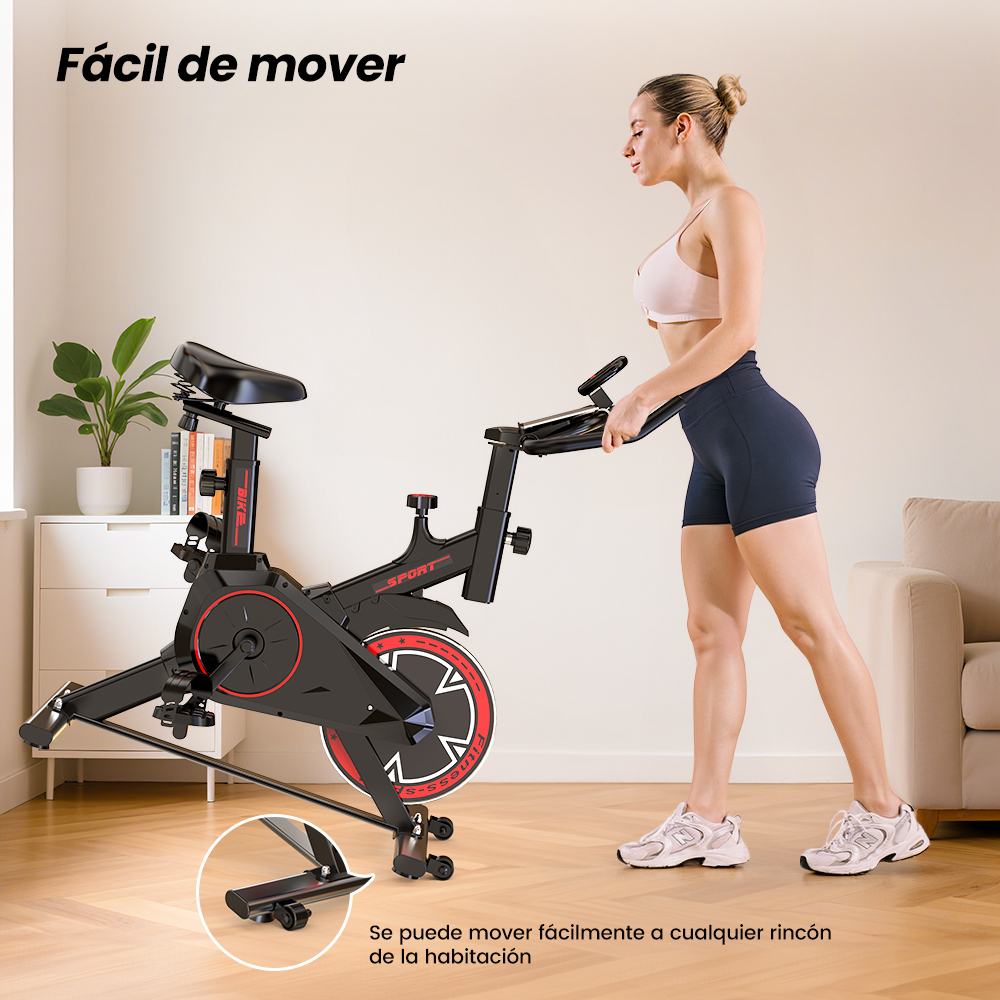 Una mujer en ropa deportiva empuja una bicicleta estática negra con detalles rojos en una habitación luminosa.