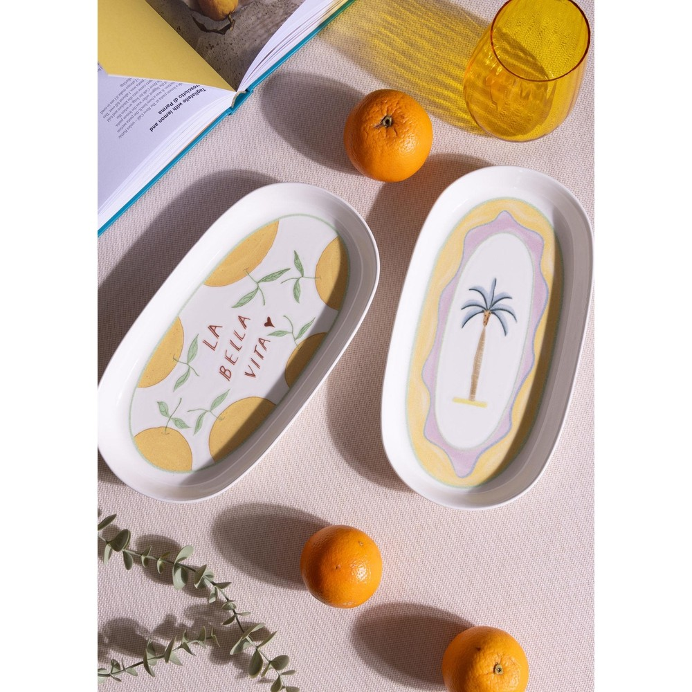 Kumaş üzerinde limon ve palmiye desenli iki oval seramik tabak, portakallar, bir dergi ve bir bardakla çevrili.