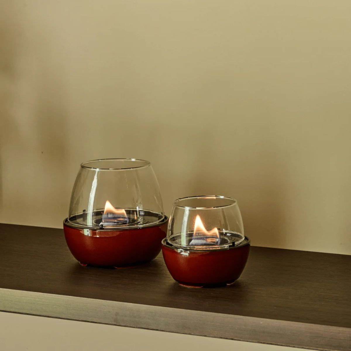 Due piccoli camini da tavolo con basi rosse e coperture di vetro bruciano su un ripiano in legno.
