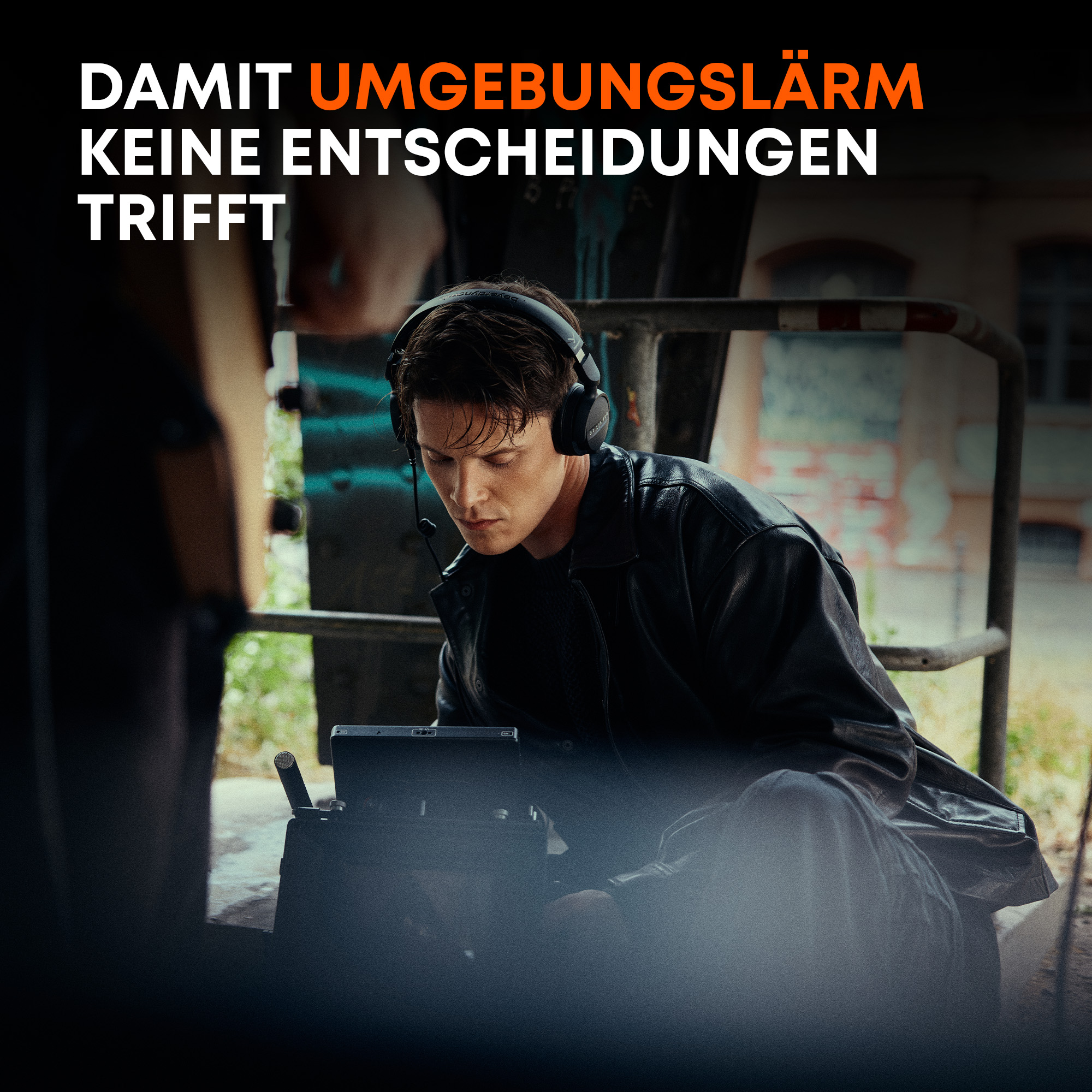 Ein Mann mit Kopfhörern und Lederjacke sitzt draußen und bedient ein elektronisches Gerät vor einer Backsteinwand.