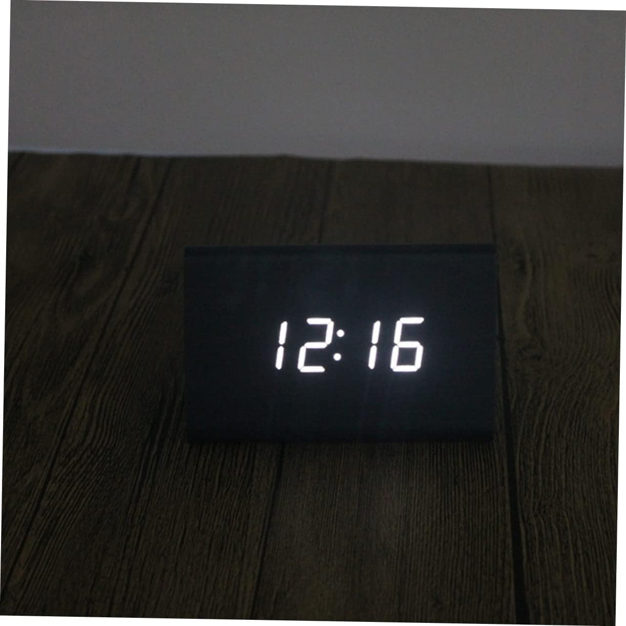 Eine schwarze Digitaluhr mit weißer LED-Anzeige 12:16 auf einer Holzoberfläche.