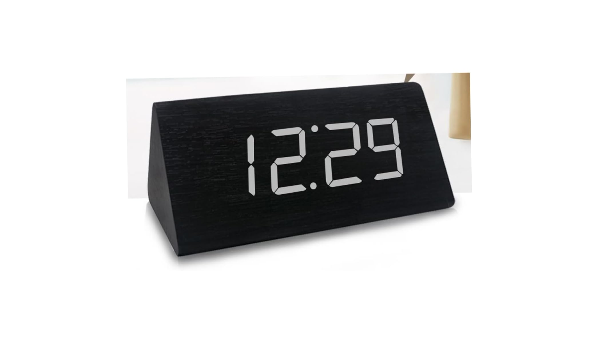 Ein dreieckiger schwarzer Holzwecker mit leuchtend weißen Digitalzahlen, die 12:29 anzeigen, auf einem hellen Tisch.