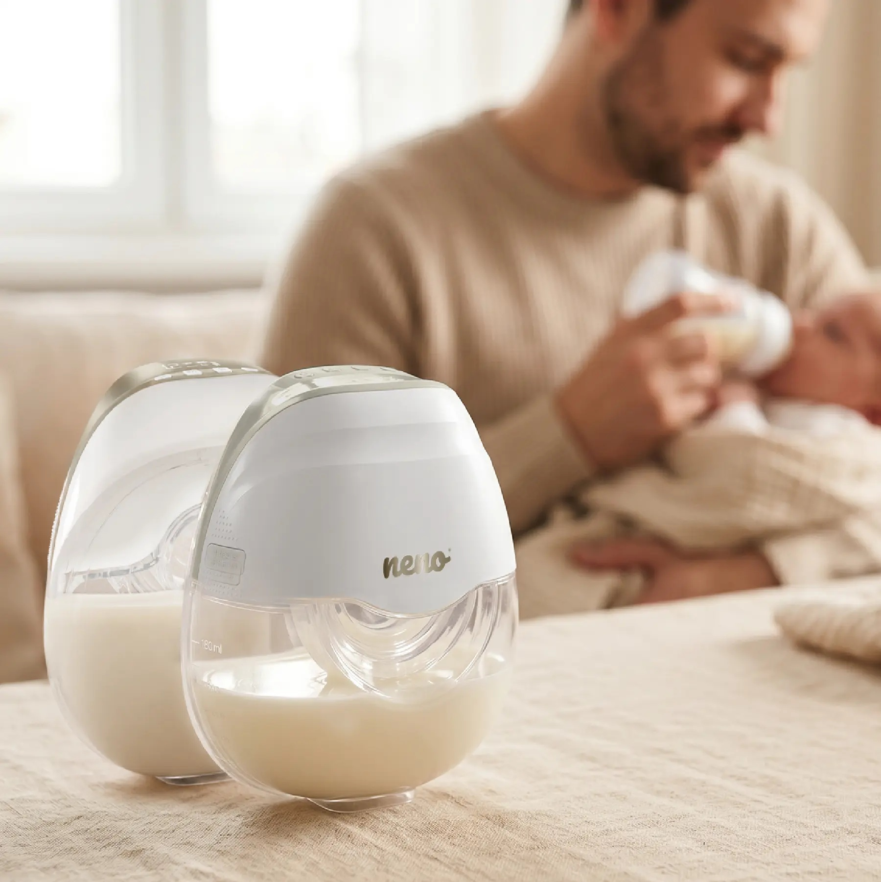 Zwei weiße Neno-Milchpumpen auf dem Tisch, im Hintergrund füttert ein Mann ein Baby.