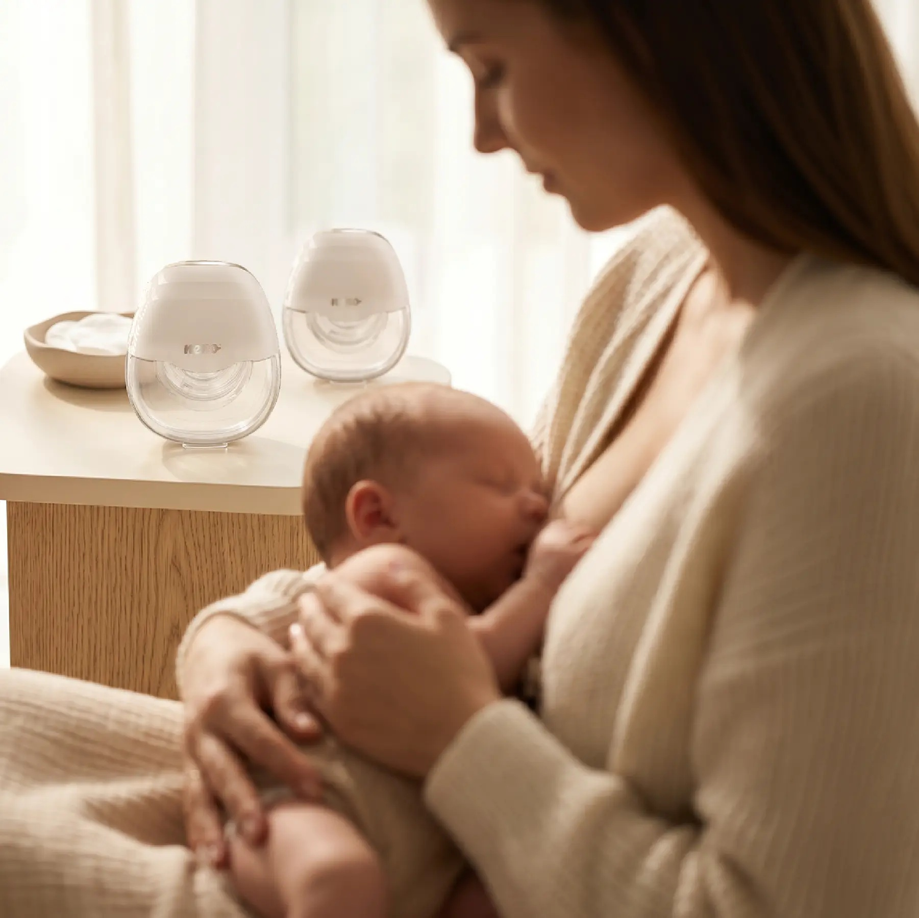 Eine Mutter hält ihr schlafendes Baby, während zwei weiße tragbare Milchpumpen auf einem Holztisch stehen.