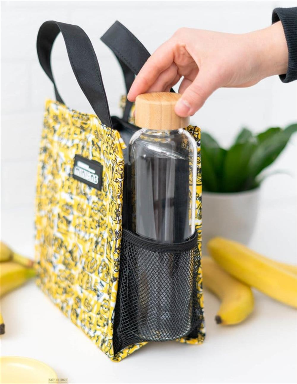 Hand legt Glasflasche mit Holzdeckel in die Netztasche einer gelben Minions-Tragetasche mit Bananen im Hintergrund.